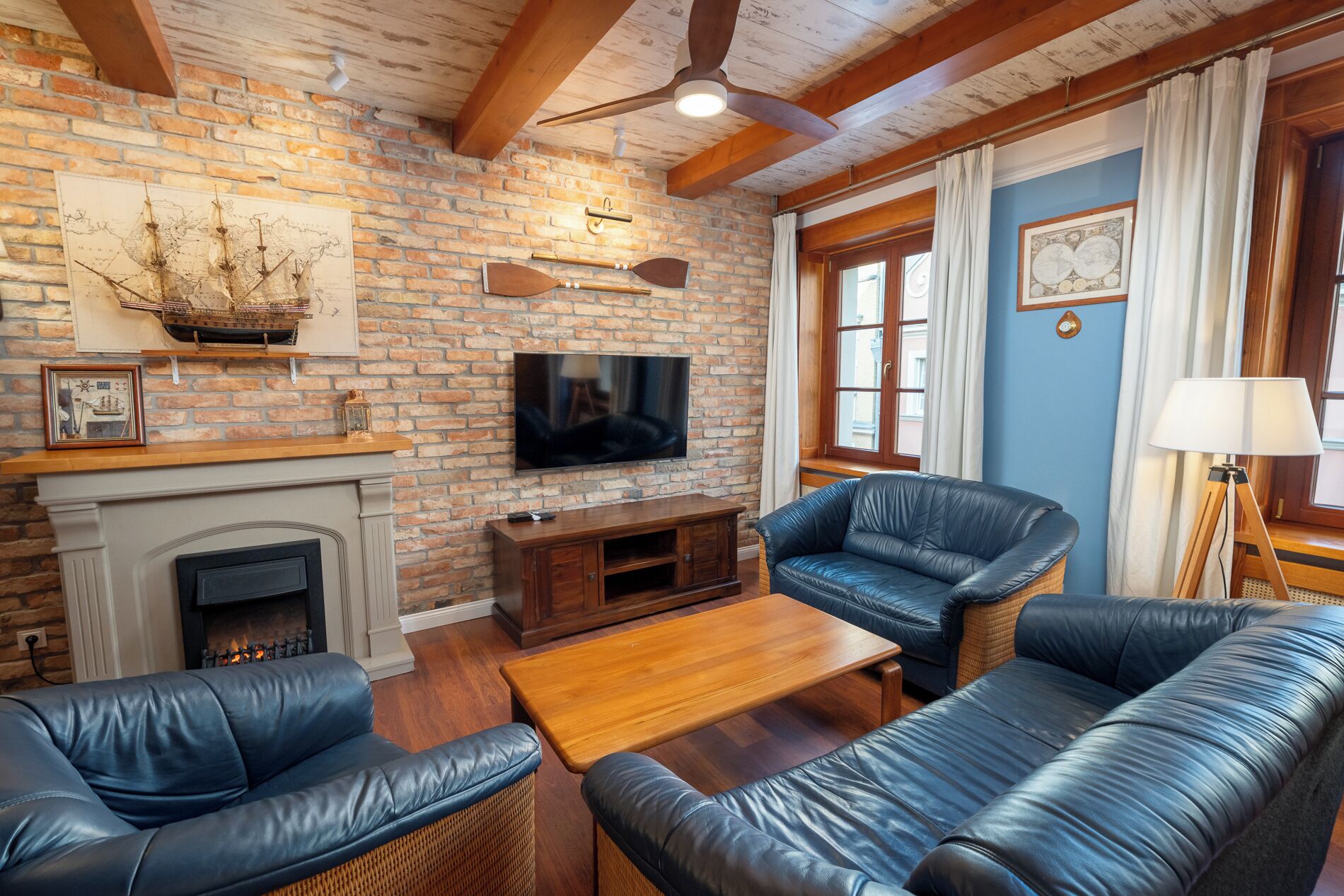 A cozy living room with a brick wall, a fireplace, leather sofas, a wooden coffee table, and a flat-screen TV.