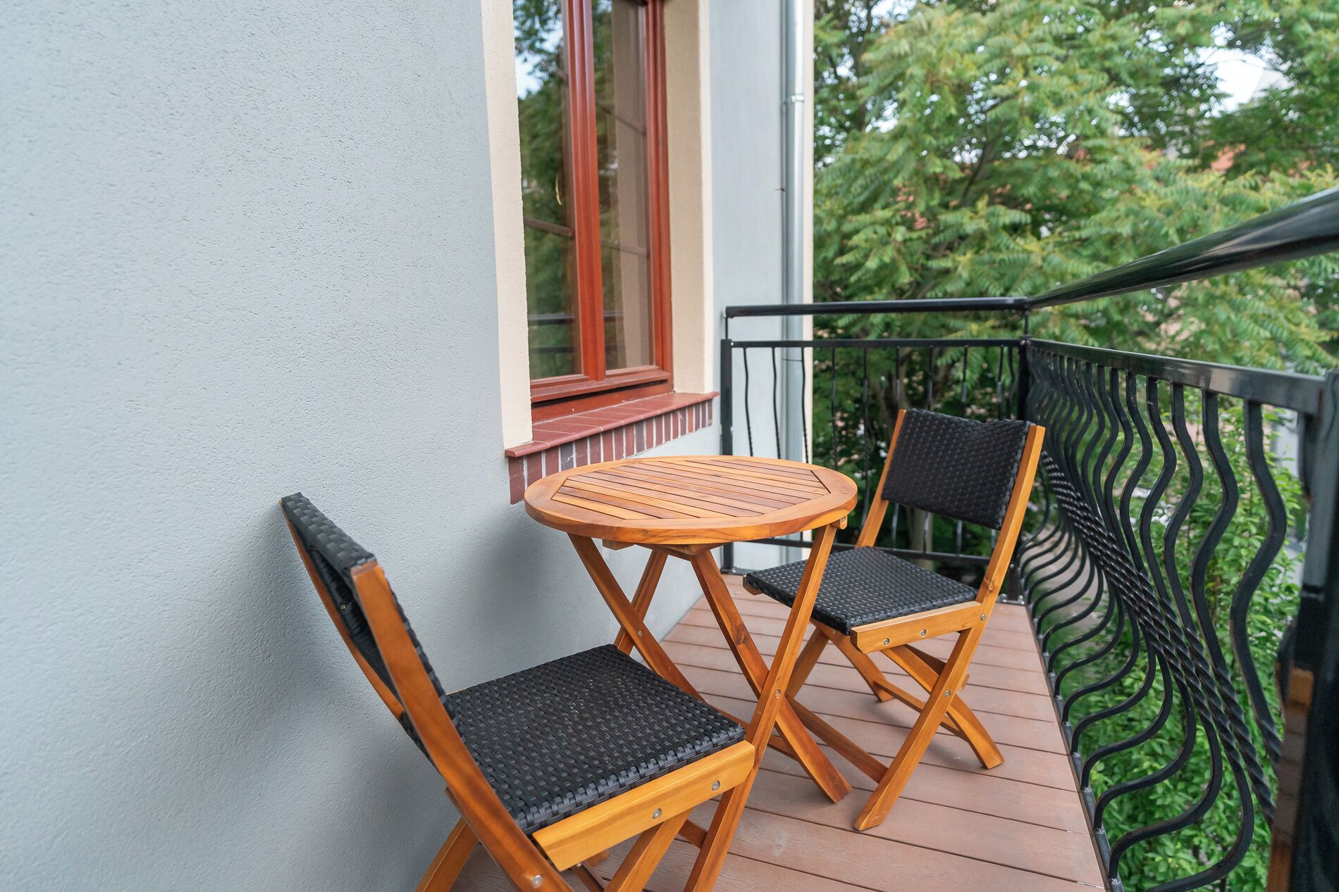 A balcony with a wooden table and two chairs, a window with a red frame, and a metal railing.