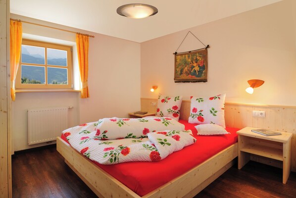 Un dormitorio con cama de madera, ropa de cama roja, una ventana con cortinas, un cuadro en la pared y lámparas de noche.