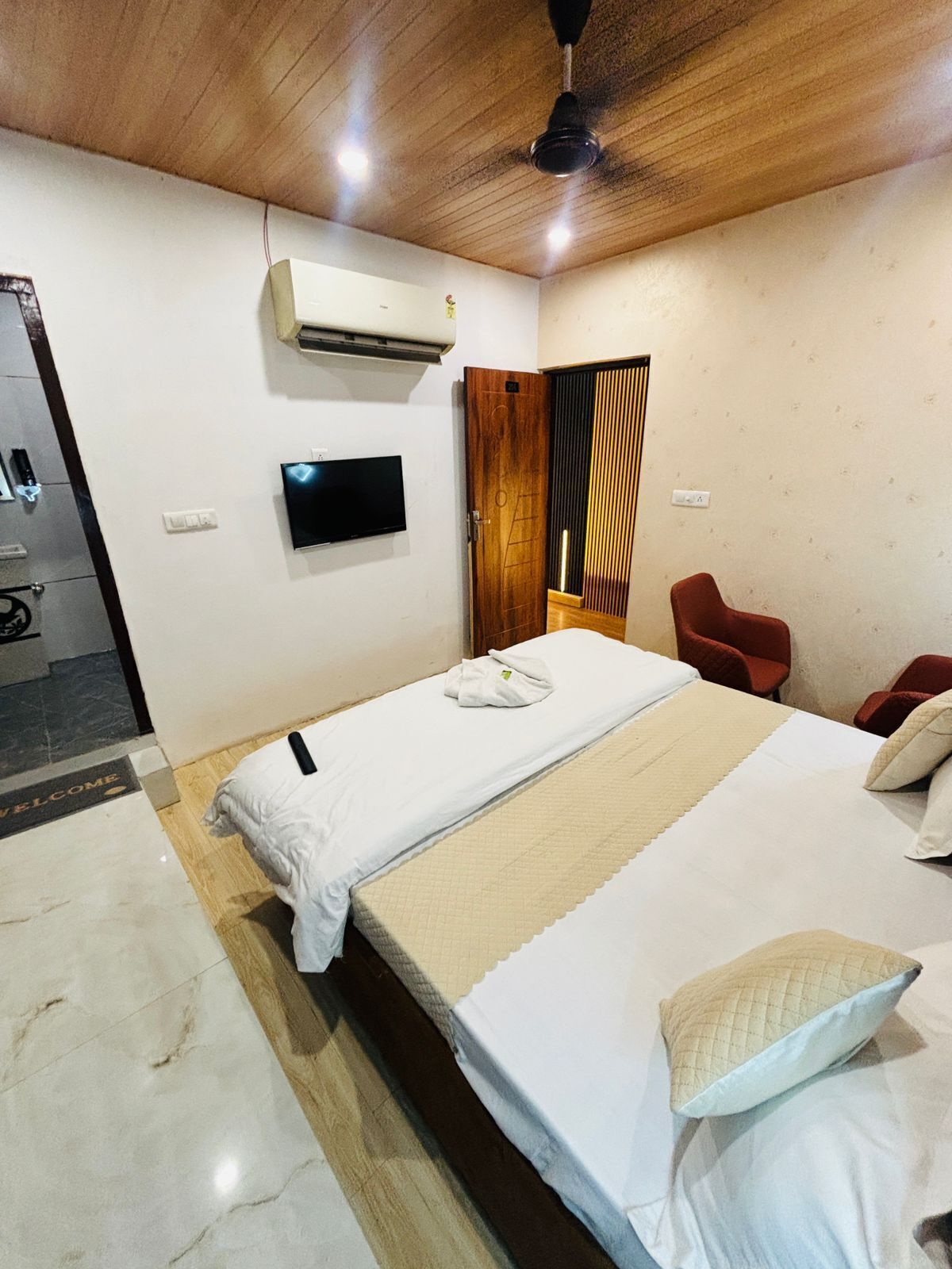 A hotel room with a bed, a TV, an air conditioner, a red chair, and a wooden ceiling.