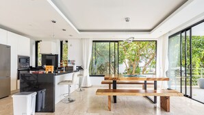 Una cocina moderna con una isla central, un área de comedor con banco de madera y grandes ventanales con vistas al verdor.