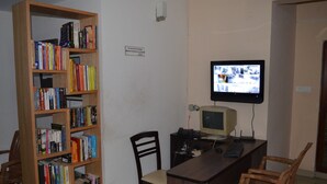 A room with a wooden bookshelf filled with books, a desk with a computer and keyboard, and a television mounted on the wall. - THE RED LOTUS GRAND (Mysore)