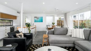 A modern living room with a grey sofa, black leather armchair, and a coffee table. There is a kitchen area with white cabinets and a dining space with a table and chairs.