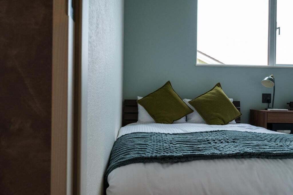 A neatly made bed with a blue blanket, two green pillows, and a bedside lamp on a wooden nightstand.