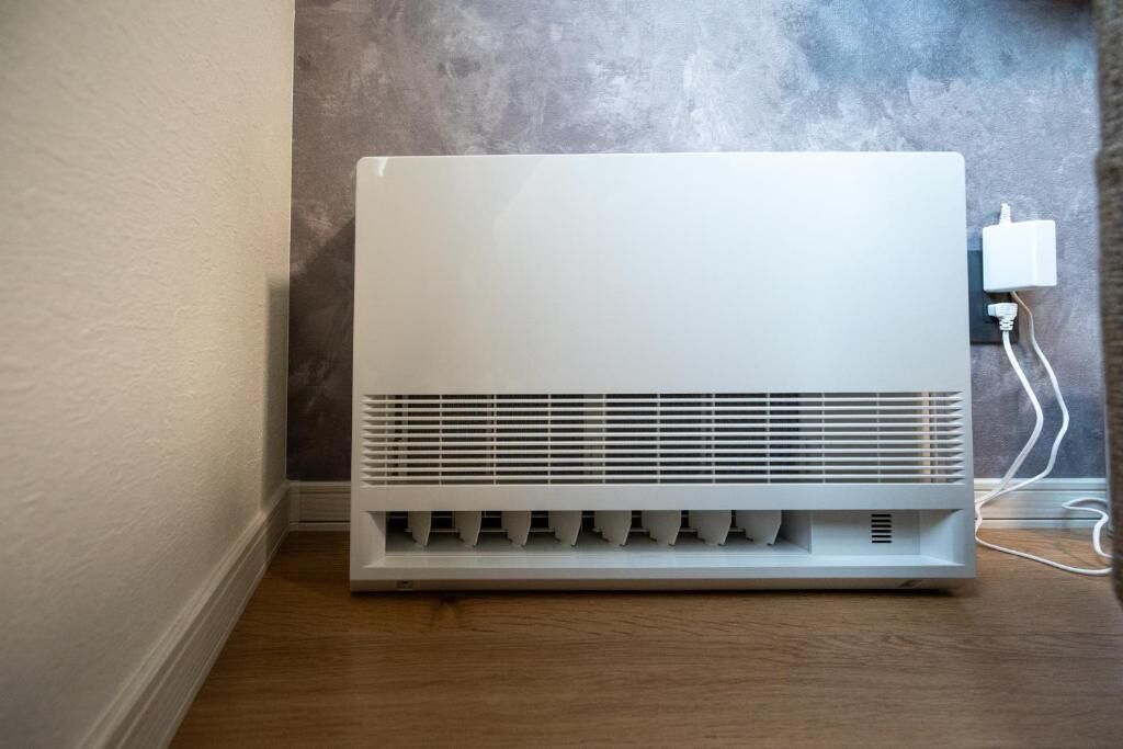 A white portable heater in a corner with a wall socket nearby.