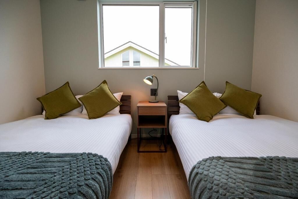 Une chambre double avec deux lits, une table de chevet, une lampe et une fenêtre donnant sur une maison.