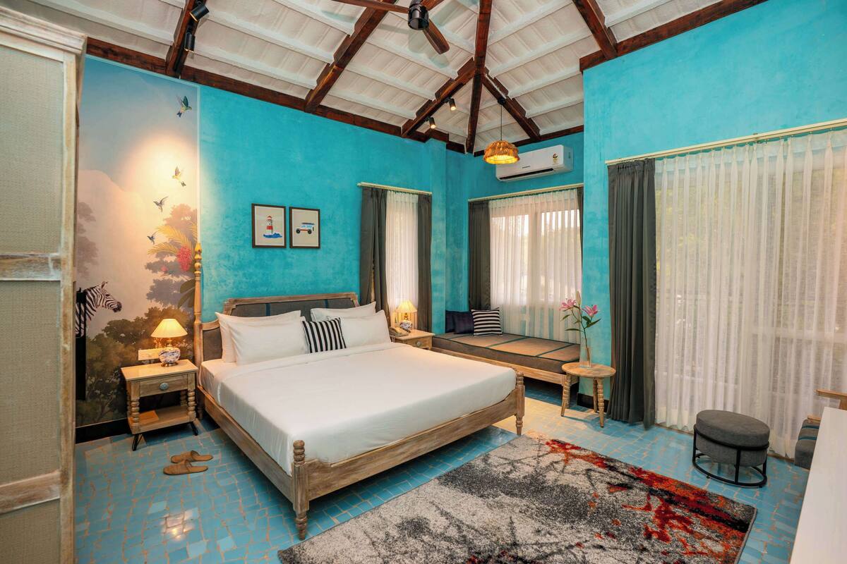 A bedroom with a large bed, a small table, a chair, a nightstand, a wall-mounted air conditioner, and a window with curtains.