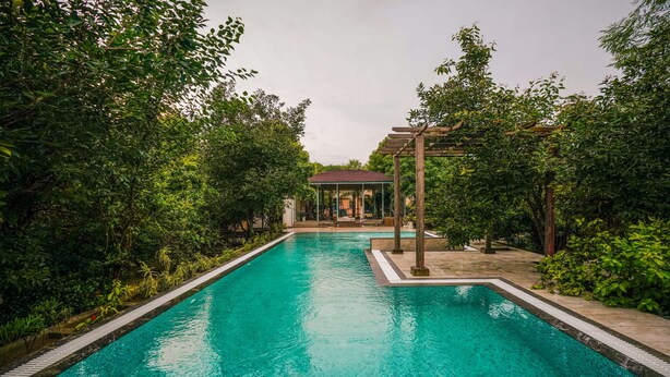 A rectangular swimming pool with clear blue water, surrounded by lush greenery and a pergola structure. - Sisodia Bagh Luxury Boutique Resort (Amer)