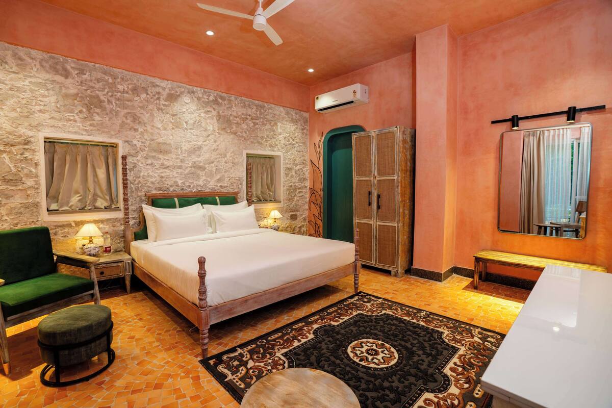 A bedroom with a bed, a green armchair, a small table with a lamp, a bench, a mirror, and a door.
