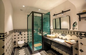 Ein Badezimmer mit einer gläsernen Duschkabine, einem Waschbecken mit Spiegel und einem Fliesenboden mit geometrischem Muster.