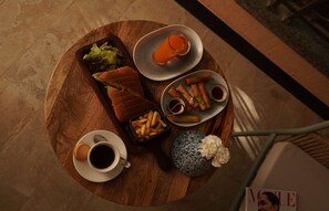 Ein runder Holztisch mit verschiedenen Speisen, darunter ein Sandwich, Pommes, Frühlingsrollen und ein Glas Orangensaft. Außerdem befinden sich zwei Kaffeetassen und eine dekorative Vase darauf.