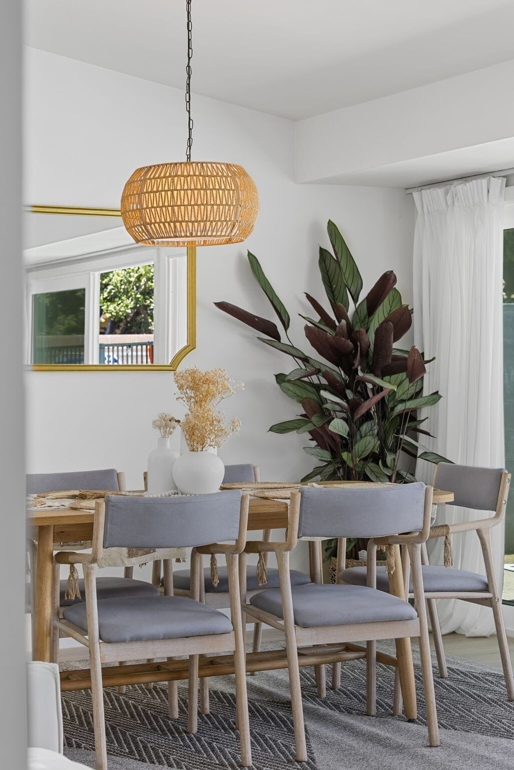 A dining area with a round table, wooden chairs, a woven pendant light, and a large plant.