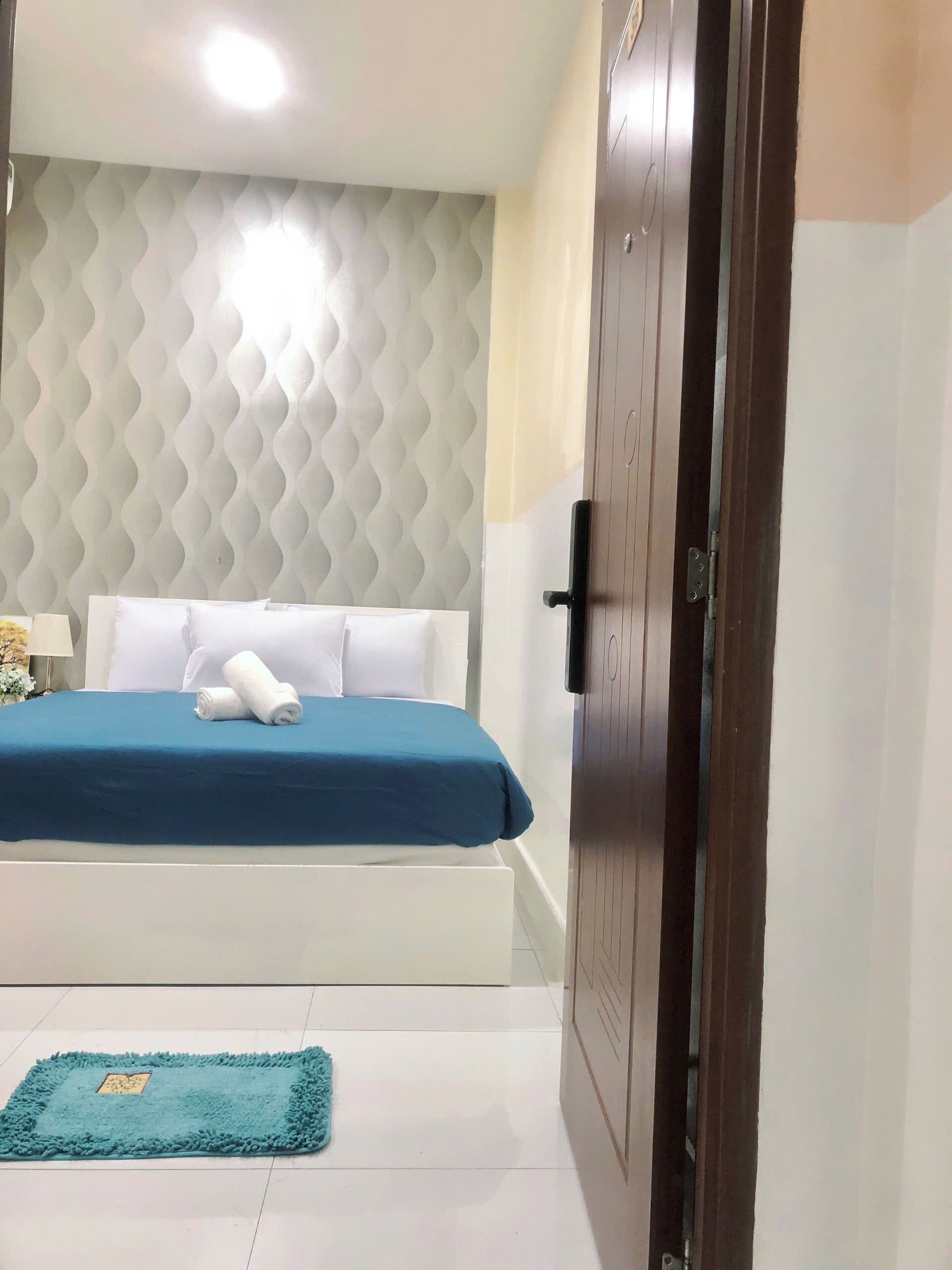 Een hotelkamer met een bed, een blauw sprei, een wit hoofdeinde, een houten deur en een blauw vloerkleed.