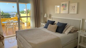 Un dormitorio con cama, mesitas de noche, lámpara y vistas a un balcón con mobiliario exterior.