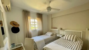 Habitación de hotel con dos camas, un escritorio, una silla, un ventilador de techo y una ventana con cortinas.