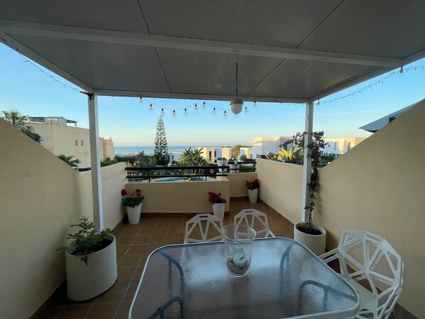 A covered outdoor patio with a glass table, white chairs, potted plants, and a view of the sea.