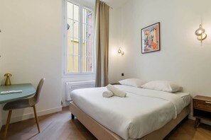 A bedroom with a bed, a desk, a chair, a window, and a wall-mounted light fixture.