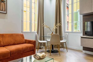 Een woonkamer met een bank, een glazen salontafel, een televisie, een eettafel met stoelen en een vaas met takken.