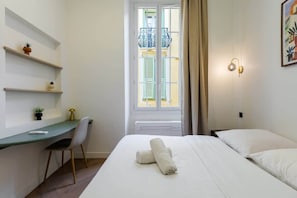 A hotel room with a bed, a desk, a chair, a window with shutters, and a wall-mounted light fixture.