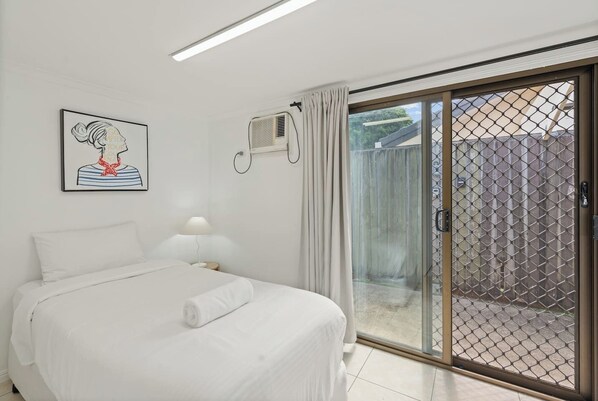 A bedroom with a bed, a wall-mounted air conditioner, a window with curtains, and a view of a fenced outdoor area.