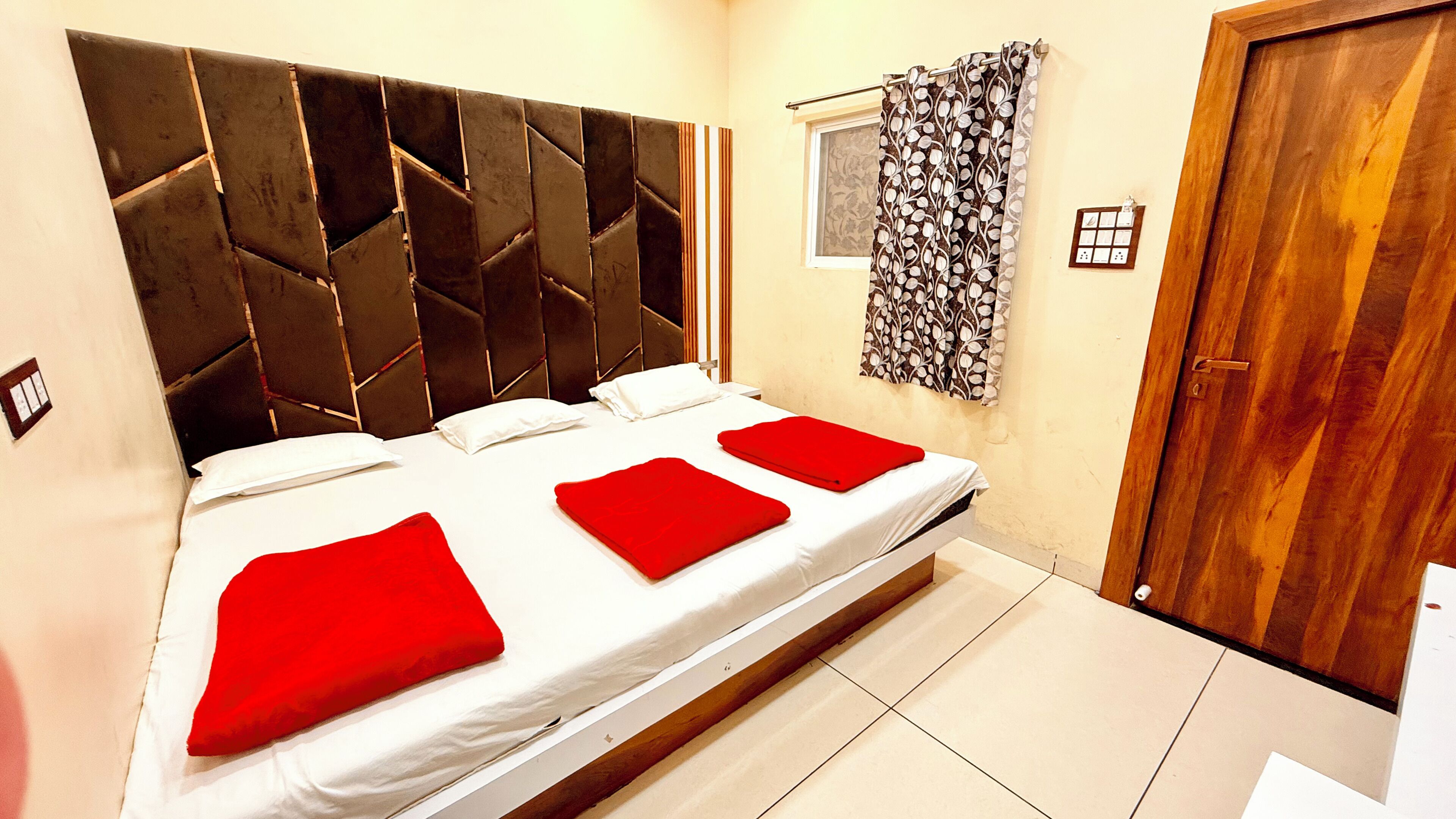 A bedroom with a modern headboard, a bed with white linens and red pillows, a window with patterned curtains, and a wooden door.