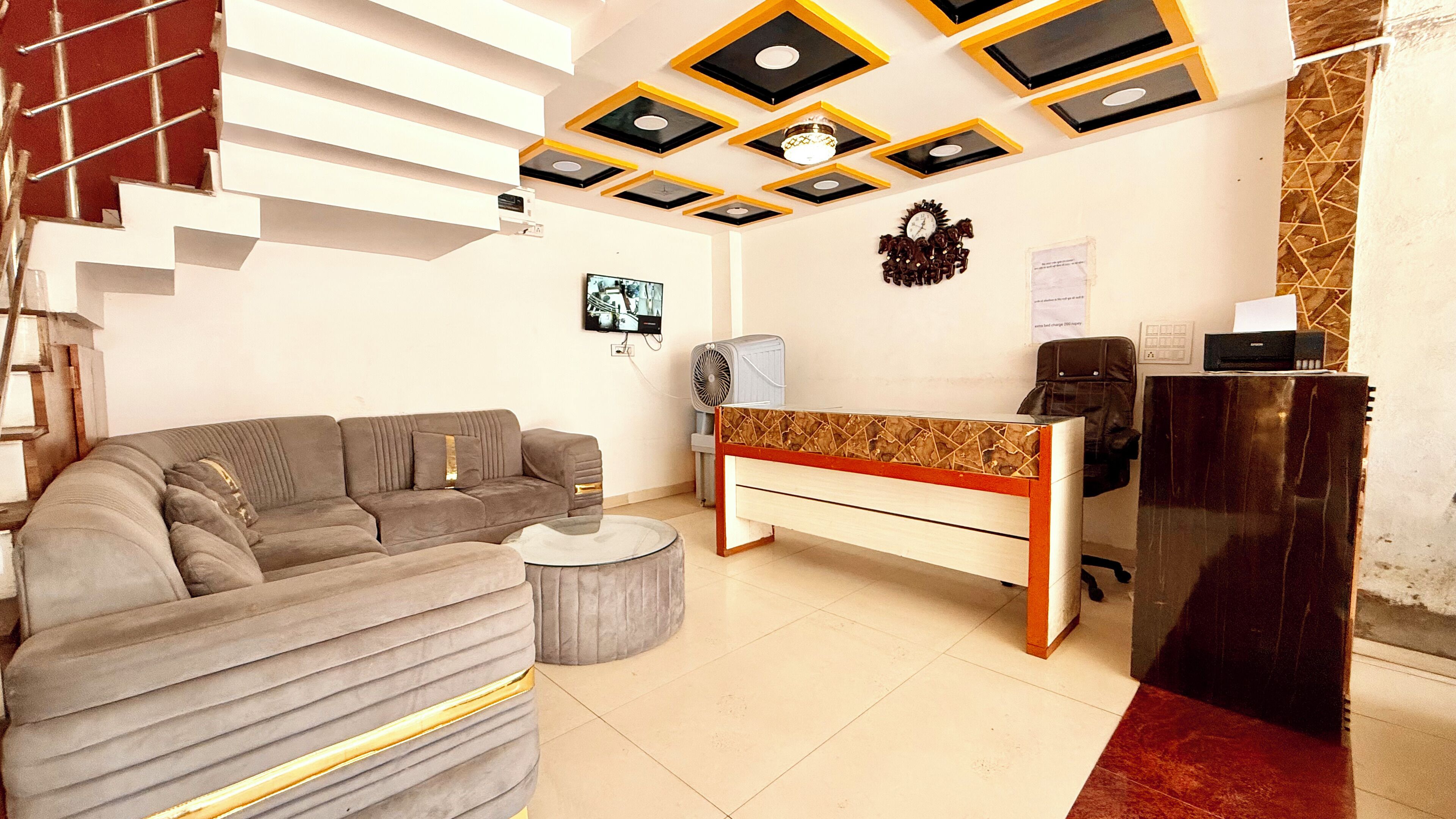 Un salon avec un canapé, une table basse, un bureau et une chaise. Un escalier se trouve à gauche et une horloge est accrochée au mur.