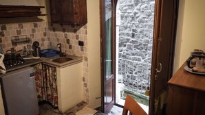 Cocina compacta con fregadero, cocina y armarios de madera. Se ve un refrigerador pequeño, y una puerta da a un balcón con muro de piedra.