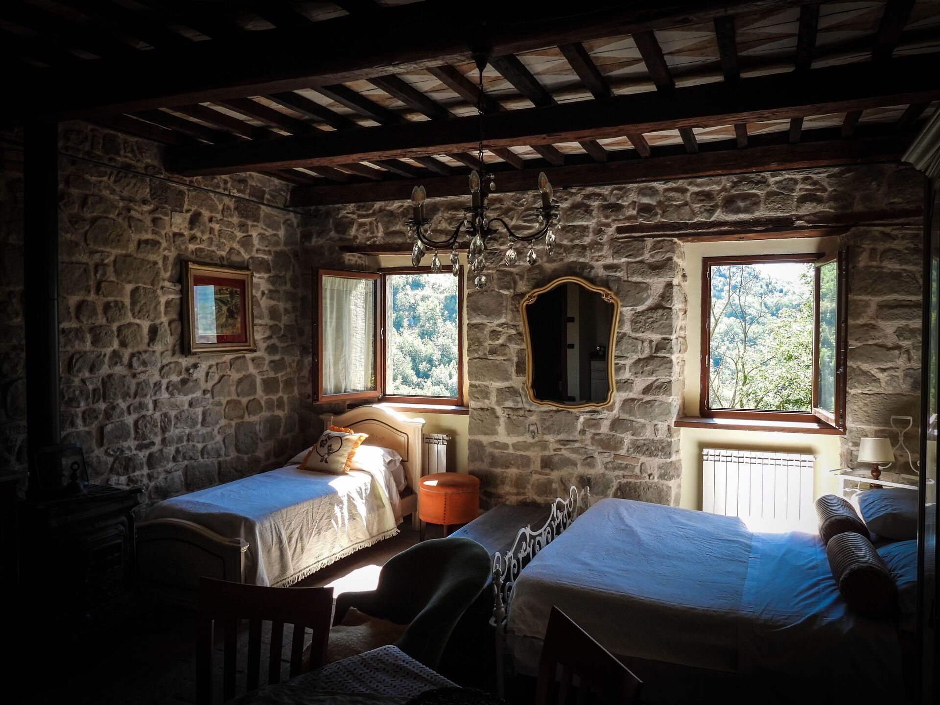 Une chambre rustique aux murs de pierre, dotée de deux lits, d’un lustre et de fenêtres offrant une vue sur la verdure.