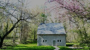A stone cottage with a small porch, surrounded by lush greenery and flowering trees.