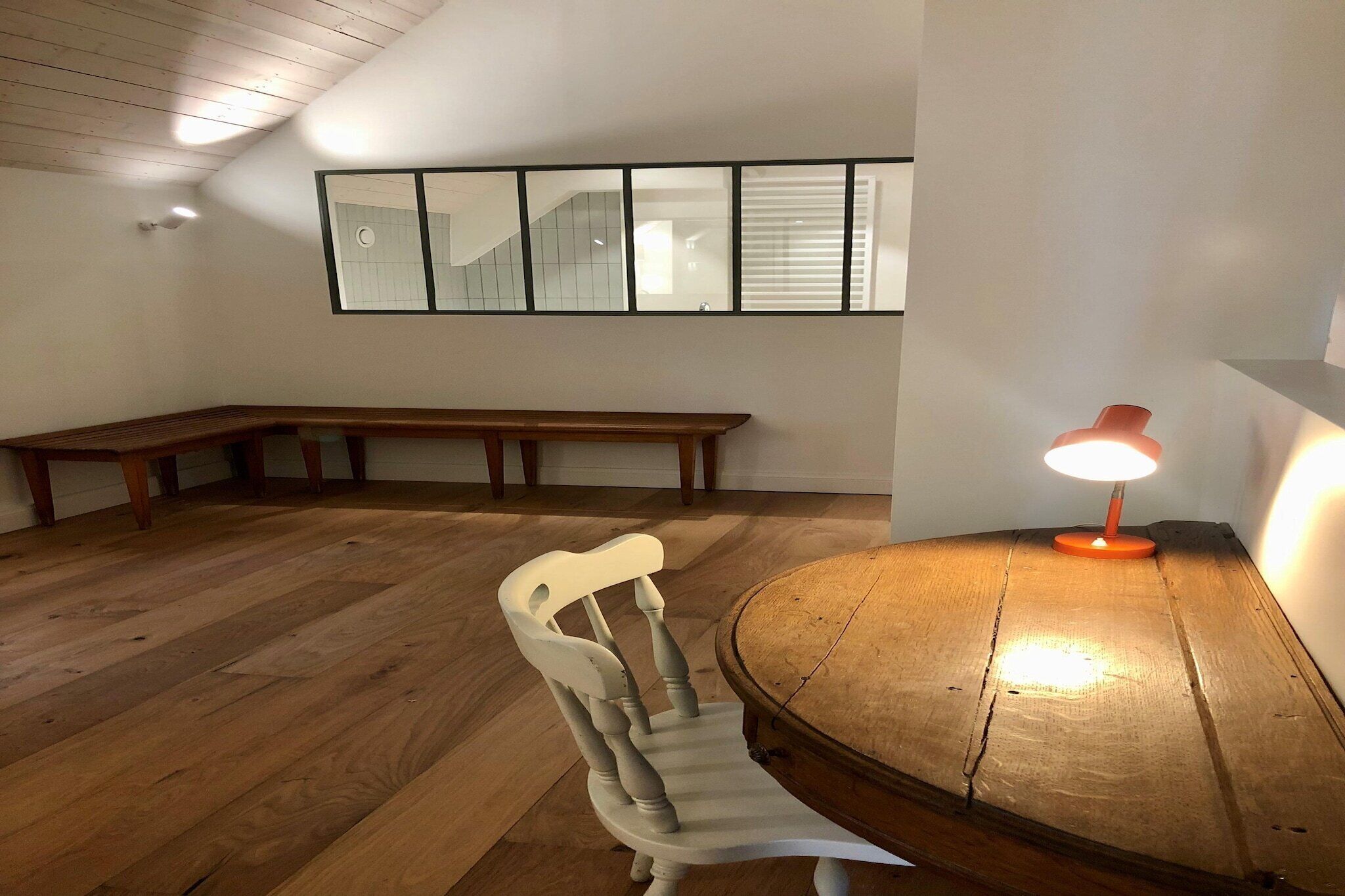 A room with a wooden floor, a wooden bench, a wooden chair, a table with a lamp, and a wall with a window and a framed picture.