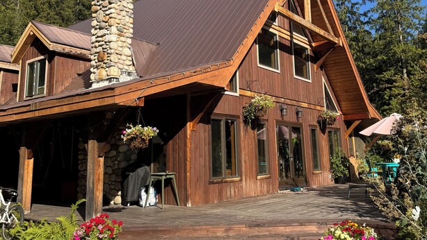 Une cabane en bois avec une cheminée en pierre, de grandes fenêtres et une véranda couverte avec des sièges d’extérieur.