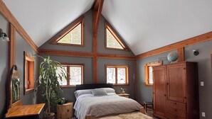 Une chambre avec un plafond en pente, des poutres en bois, un lit recouvert d’un plaid en fourrure, une commode en bois et une grande fenêtre donnant sur la forêt.