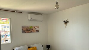 A neatly made bed with a white and yellow bedspread, a single white pillow, and a rolled white towel. There is a small bedside table with a black lamp and a wall-mounted air conditioner above it.