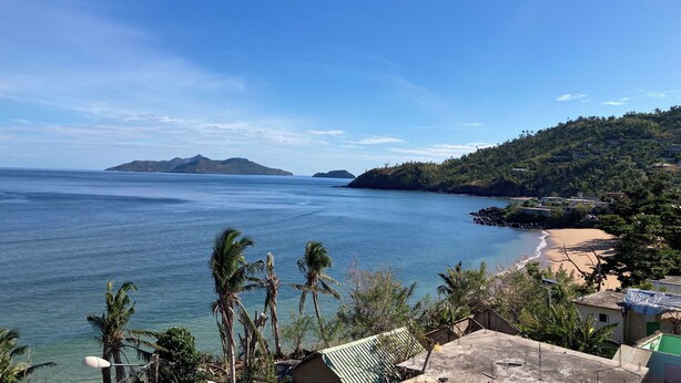 A coastal view with a beach, palm trees, and a hilly landscape.