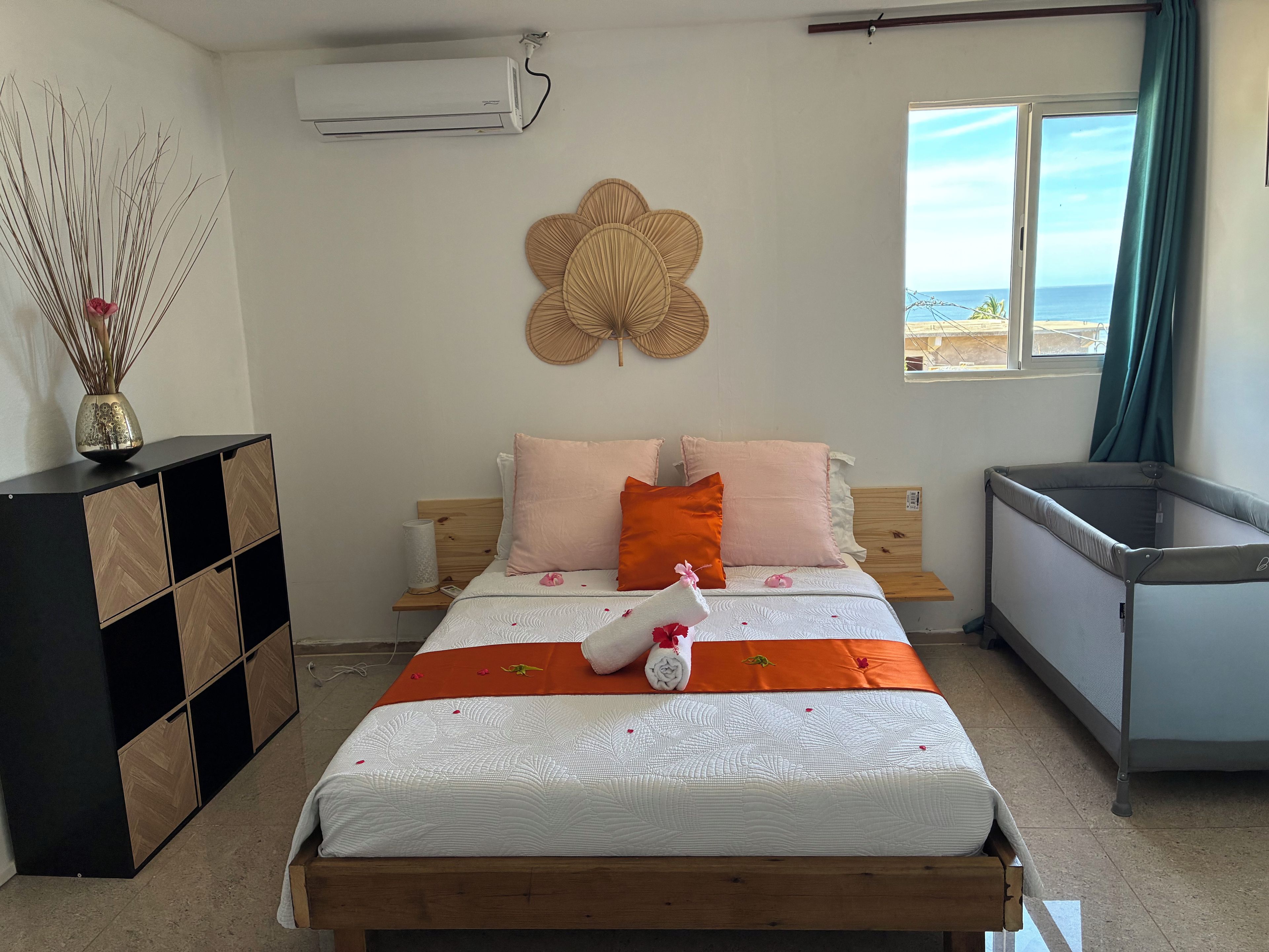 A bedroom with a bed, bedside table, air conditioning unit, decorative wall piece, and a view of the sea through a window.