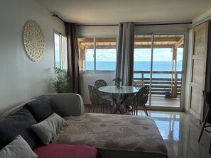 A room with a bed, a dining table with chairs, a view of the sea, and a balcony with a railing. - T2 cosy Côte îlots au bord de la plage vue panoramique, coucher de soleil. (Canton d'Acoua)
