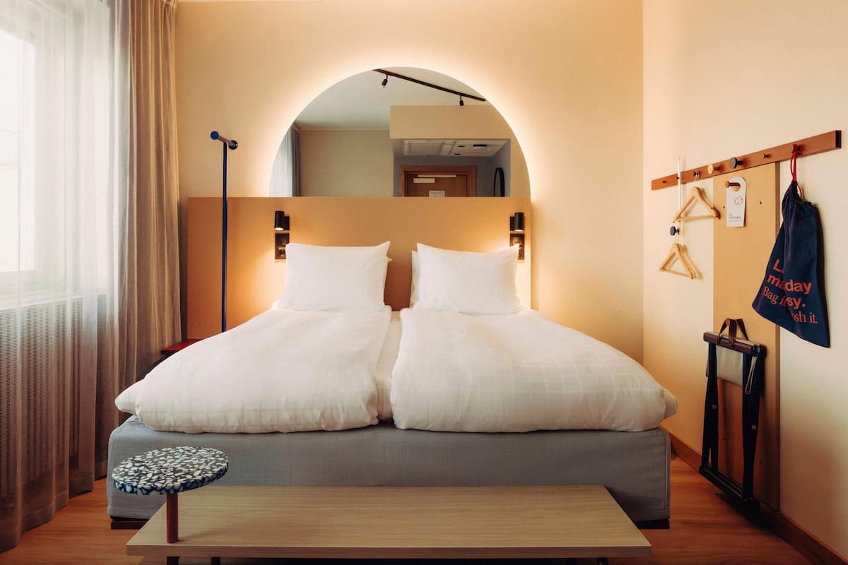 A double bed with white bedding, a small round table, and a bench.