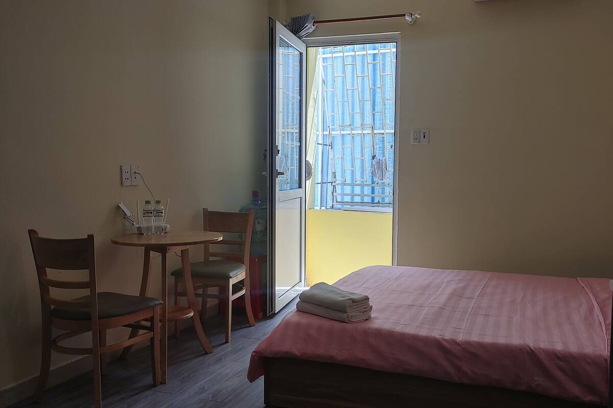 A small, simple room with a bed, a table with chairs, and a window with a view of a blue building.