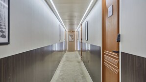 Ein langer, schmaler Korridor mit Teppichboden, gesäumt von Wänden mit gerahmten Bildern und einer Holztür auf der rechten Seite.