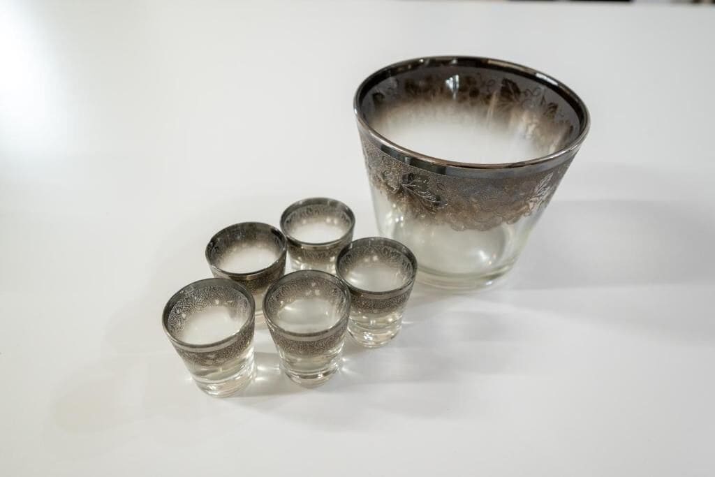 Una taza de vidrio transparente con diseño floral y cuatro pequeños vasos para chupitos a su lado.