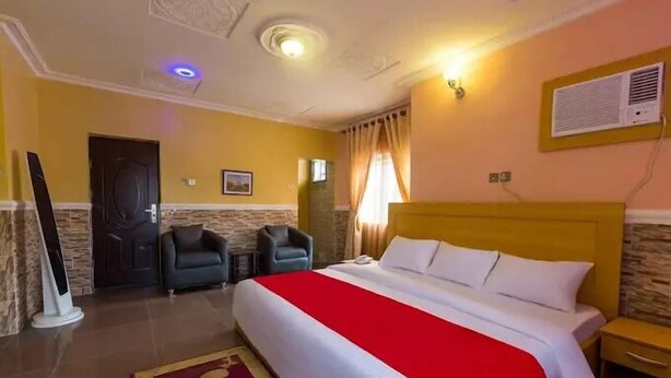 A hotel room with a bed, a nightstand, a chair, a desk, a wall-mounted air conditioner, and a door. - Casalavoro Residence (Abuja)