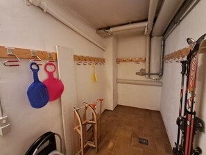 Ein schmaler Flur mit einem Holzstuhl, einem an der Wand befestigten Hakenständer und einer Reihe von Skiern, die an der Wand lehnen.