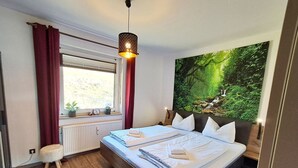 A bedroom with a bed, a window with curtains, a radiator, a small stool, and a large forest-themed wall hanging.