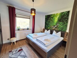 A bedroom with a bed, a window with curtains, a radiator, a small stool, and a large forest-themed wall hanging.