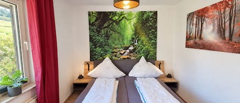 Una cama doble con cabecera, una ventana con cortinas rojas, una lámpara de pared y un cuadro con motivos forestales.