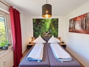 A double bed with a headboard, a window with red curtains, a wall-mounted lamp, and a forest-themed painting.