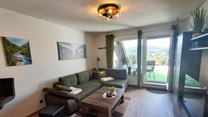 A modern living room with a sofa, a coffee table, and a TV. There are two paintings on the wall, a window with curtains, and a balcony with a chair.
