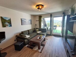 A modern living room with a sofa, a coffee table, and a TV. There are two paintings on the wall, a window with curtains, and a balcony with a chair.