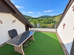 A balcony with a table and chairs, a view of a green landscape, and decorative wall elements.
