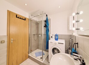 A modern bathroom with a glass-enclosed shower, a washing machine, a sink, and a mirror with lights.